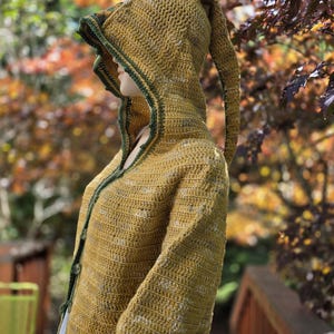 May include: A mustard yellow crochet hooded cardigan featuring a long, pointed pixie-style hood. The garment is constructed with a textured stitch pattern and finished with a dark green crocheted trim along the front opening and hood edge.