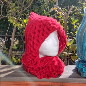 May include: A red crocheted hooded scarf with a scale pattern. The scarf is displayed on a wooden railing.