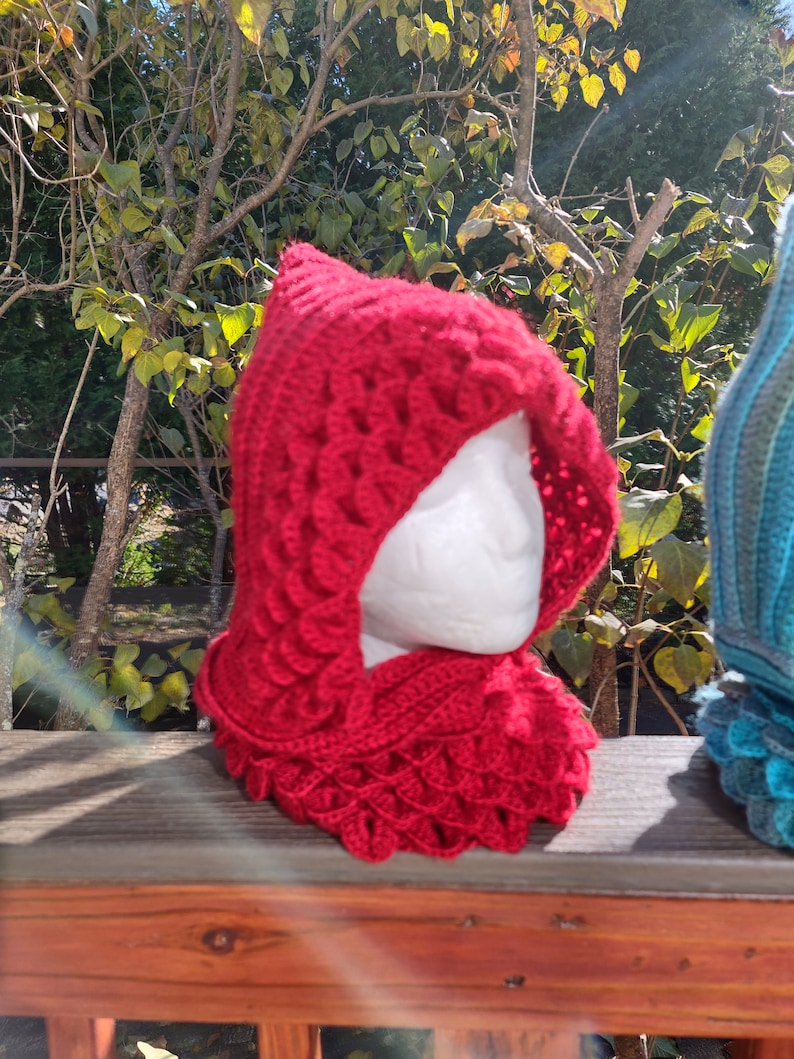May include: A red crocheted hooded scarf with a scale pattern. The scarf is displayed on a wooden railing.