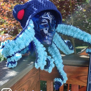 May include: A blue and light blue crocheted octopus hat with a red pom-pom on top. The hat is worn on a blue mannequin head. The hat is on a wooden railing with the text "itsinthebagcrochet" on the railing.