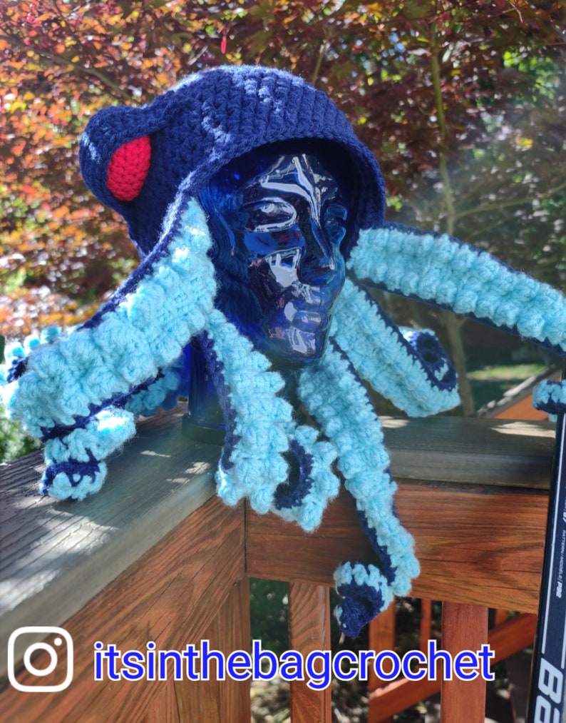 May include: A blue and light blue crocheted octopus hat with a red pom-pom on top. The hat is worn on a blue mannequin head. The hat is on a wooden railing with the text "itsinthebagcrochet" on the railing.