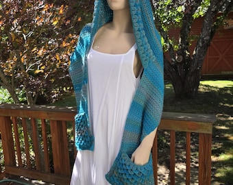 May include: A hooded scarf in shades of blue and grey with a textured scale pattern. The scarf features two integrated pockets at the ends, each displaying the same scale-like stitch detail. The garment is draped over a plain white sleeveless dress.