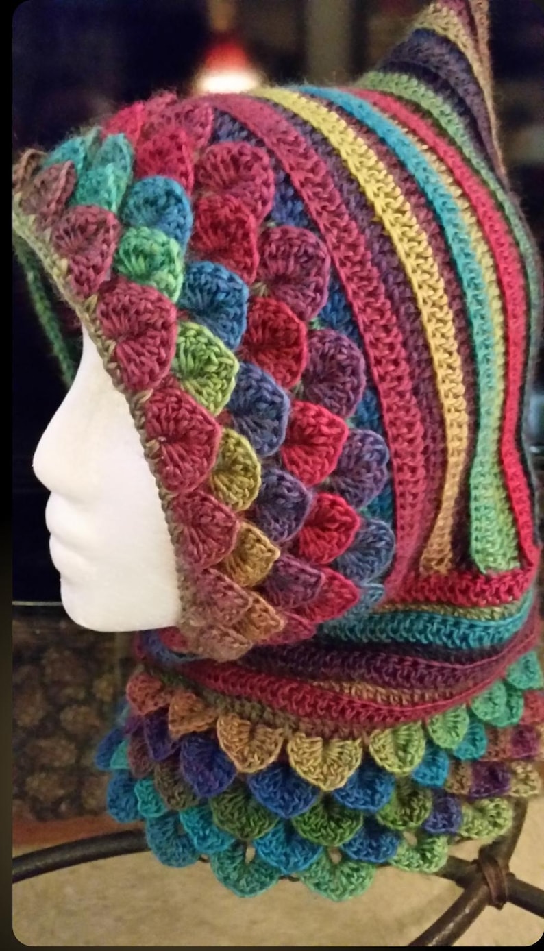 May include: A colorful crocheted hooded cowl with a rainbow striped pattern and a textured, scale-like design. The cowl is made with a variety of yarn colors, including red, orange, yellow, green, blue, purple, and brown.