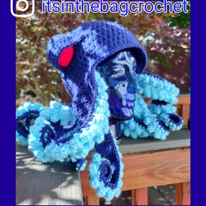 May include: A blue and light blue crocheted octopus hat with a red pom-pom on top. The hat is on a mannequin head and is sitting on a wooden surface. The Instagram handle "itsinthebagcrochet" is visible in the top right corner of the image.