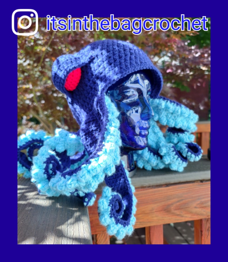 May include: A blue and light blue crocheted octopus hat with a red pom-pom on top. The hat is on a mannequin head and is sitting on a wooden surface. The Instagram handle "itsinthebagcrochet" is visible in the top right corner of the image.