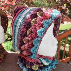 May include: A colorful crocheted hooded cowl with a pointed top and a scalloped edge. The cowl is made in shades of teal, purple, brown, and pink.