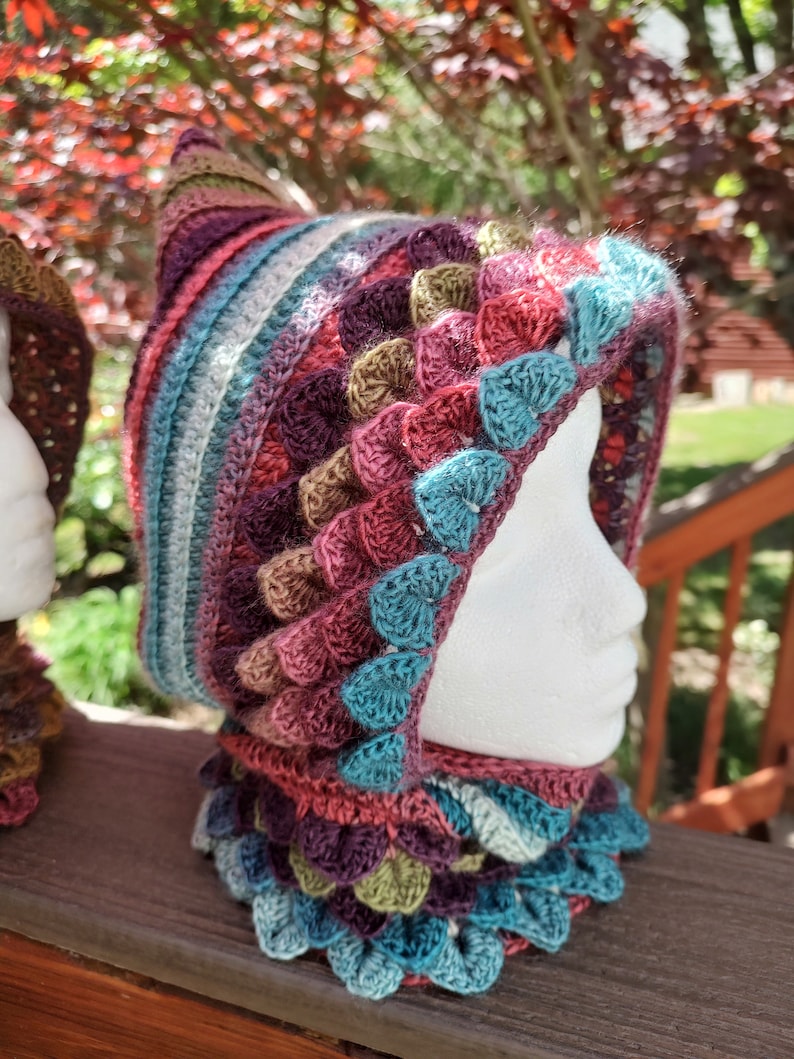 May include: A colorful crocheted hooded cowl with a pointed top and a scalloped edge. The cowl is made in shades of teal, purple, brown, and pink.