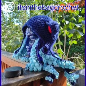 May include: Blue and turquoise crocheted octopus hat with a red pom-pom on top. The hat is on a mannequin head and is sitting on a wooden railing. The text "itsinthebagcrochet" is visible in the top right corner of the image.