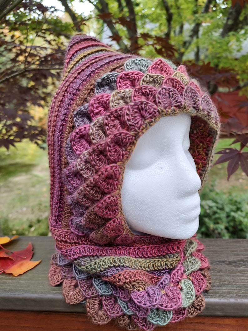 May include: A crocheted hooded cowl with a textured, scale-like pattern in shades of pink, brown, green, and purple.