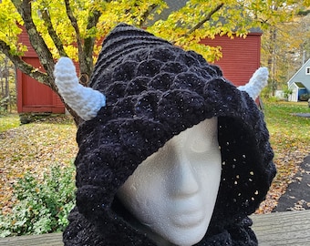 May include: A black crocheted dragon hood with white horns. The hood is designed with a scale pattern and a built-in scarf. The hood is displayed on a white mannequin head, set against a backdrop of fall foliage.