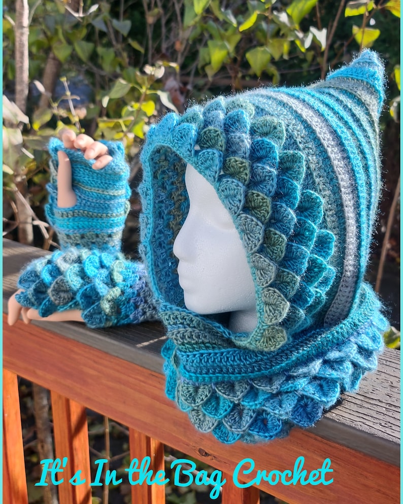 May include: A blue and green crocheted hooded cowl with matching fingerless gloves. The cowl has a pointed hood and a textured, scale-like pattern. The text "It's In the Bag Crochet" is visible at the bottom of the image.
