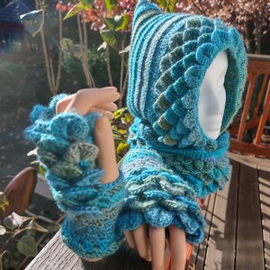 May include: A teal blue and green crocheted hooded cowl with a dragon scale pattern. The cowl has a pointed hood and matching fingerless gloves.