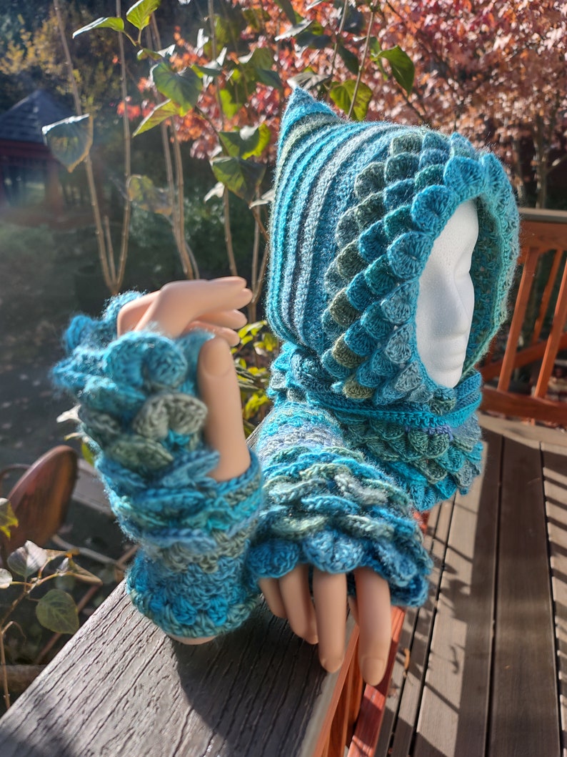 May include: A teal blue and green crocheted hooded cowl with a dragon scale pattern. The cowl has a pointed hood and matching fingerless gloves.