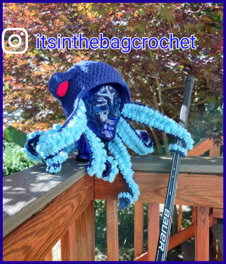 May include: A blue and light blue crocheted octopus hat with a red pom-pom on top. The hat is sitting on a wooden railing with a hockey stick leaning against it. The Instagram handle "itsinthebagcrochet" is visible in the top left corner of the image.
