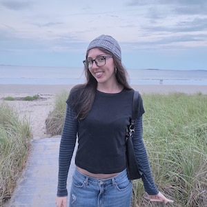 May include: A grey crocheted beanie, a black short-sleeved t-shirt layered over a grey and black striped long-sleeved shirt, blue denim jeans, and a black shoulder bag. The outfit is completed with black-rimmed glasses.