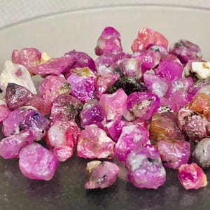 May include: A collection of raw ruby gemstones in various shades of pink and red. The rough-cut stones have a natural, unpolished appearance, with some showing hints of white and black. The gemstones are scattered, creating a textured and colorful display.
