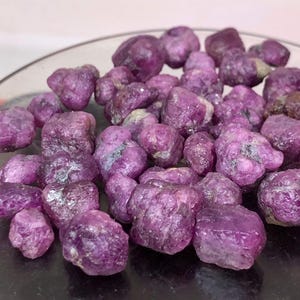 Puede incluir: Una colección de piedras preciosas de rubí en bruto y sin pulir en varios tonos de magenta intenso y púrpura. Estos pequeños especímenes minerales irregulares presentan una superficie cristalina texturizada con facetas naturales y cualidades opacas.