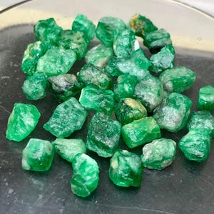 May include: A collection of small, raw emerald gemstones with a vibrant green colour and irregular, jagged shapes. The stones exhibit varying degrees of translucency and natural surface textures, scattered across a smooth, dark, reflective surface.