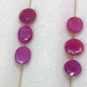 Peut inclure: Six rubis à facettes de forme ovale disposés en deux colonnes verticales. Les pierres précieuses présentent une couleur rouge magenta vive et profonde avec des facettes géométriques visibles qui reflètent la lumière sur leurs surfaces.