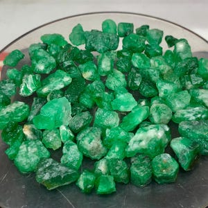 Puede incluir: Una colección de pequeñas piedras preciosas de esmeralda en bruto en varios tonos de verde vibrante. Las piedras presentan formas irregulares y dentadas con texturas translúcidas y opacas, mostrando estructuras cristalinas naturales dispersas sobre una superficie circular.