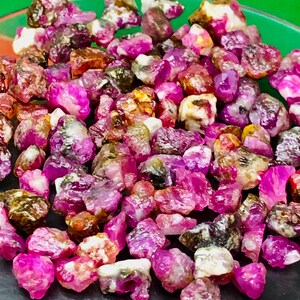 Op de afbeelding: Een verzameling ruwe robijn edelstenen in verschillende tinten roze, rood en bruin. De ongeslepen stenen worden getoond op een donkere ondergrond, waarbij hun natuurlijke, onregelmatige vormen en kristallijne structuren zichtbaar zijn.