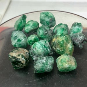 以下が含まれることがあります： 暗い表面に展示された、さまざまなサイズと形状の生の緑色のエメラルド宝石のコレクション。石は、鮮やかなものから深いものまで、さまざまな緑の色合いを示し、一部には自然なインクルージョンとテクスチャが見られます。宝石は研磨されていません。