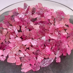 May include: A collection of small, raw, unpolished pink and magenta tourmaline gemstone fragments. The stones exhibit varying degrees of translucency, jagged edges, and natural crystalline textures, scattered across a smooth, reflective surface.