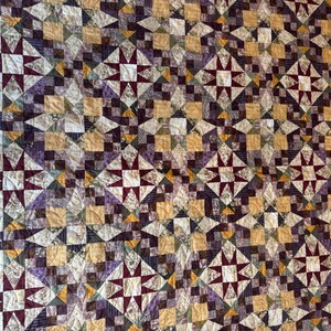 May include: A quilt with a patchwork design featuring a variety of colors, including brown, purple, and gold. The quilt is made up of squares and triangles, and it has a starburst pattern in the center of each square.