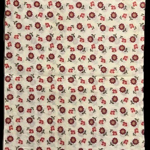 May include: Cream-colored fabric with a repeating pattern of burgundy flowers and berries. The flowers have a dark center and are surrounded by petals. The berries are small and clustered. The edge of the fabric has a green floral trim.
