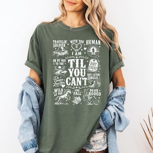 May include: A moss green t-shirt featuring white graphic text and illustrations including a heart, a horse, a cowboy boot, and the phrases Travelin' Soldier, Human, Dirt Cheap, She's Acting Single, Wild As You, The Fall, and Dear Rodeo.