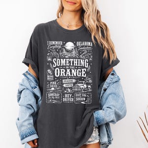 May include: A charcoal grey short-sleeve t-shirt featuring a white graphic print with the text "Something in the Orange" and various illustrations including a truck, a moon, and desert landscapes, layered under a light blue denim jacket.