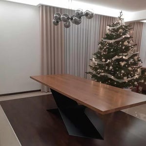 Może przedstawiać: Nowoczesny drewniany stół jadalny z jasnobrązową powierzchnią i czarną geometryczną podstawą. Stół znajduje się w pokoju z choinką ozdobioną światełkami i ozdobami. Pokój ma neutralne zasłony i nowoczesny żyrandol.