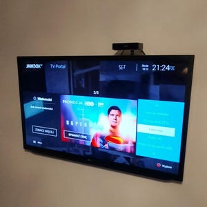 Può includere: Un televisore a schermo piatto montato a parete che mostra un'interfaccia del portale TV. Lo schermo mostra le informazioni sul programma, incluso il testo "SUPERMAN" e "PROMOCJA HBO." Un piccolo dispositivo nero è in cima.