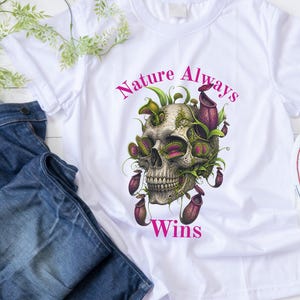 Puede incluir: Una camiseta blanca con un gráfico de un cráneo humano entrelazado con plantas carnívoras moradas y verdes, incluyendo venus atrapamoscas y plantas jarro. El texto "Nature Always Wins" está impreso en rosa sobre y debajo del gráfico.