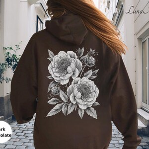 K&ouml;nnte beinhalten: Dunkelbrauner Kapuzenpullover mit detailliertem schwarz-wei&szlig;em Blumendesign auf dem R&uuml;cken. Der Hoodie hat eine Kapuze mit Kordelzug und eine K&auml;ngurutasche. Der Text "Dark Chocolate" befindet sich unten links.