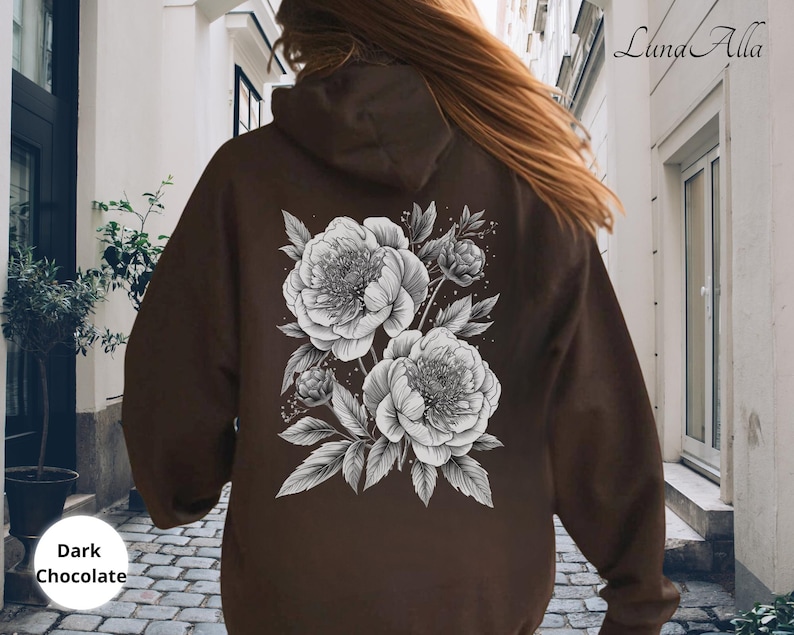 K&ouml;nnte beinhalten: Dunkelbrauner Kapuzenpullover mit detailliertem schwarz-wei&szlig;em Blumendesign auf dem R&uuml;cken. Der Hoodie hat eine Kapuze mit Kordelzug und eine K&auml;ngurutasche. Der Text "Dark Chocolate" befindet sich unten links.