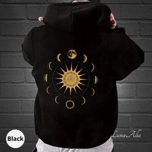 Puede incluir: Sudadera con capucha negra con un diseño dorado de sol y fases lunares en la espalda. El diseño incluye un sol central con lunas crecientes circundantes y una luna llena en la parte superior. La palabra "Black" está en un círculo blanco en la parte inferior izquierda.