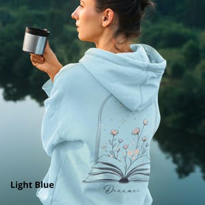 May include: A light blue hooded sweatshirt featuring a graphic on the back of an open book with flowers growing out of the pages inside an arched frame, accompanied by the word Dreamer in cursive script below the illustration.