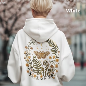 Op de afbeelding: Een witte hoodie met een grote afbeelding op de achterkant van een mot in een cirkel van varens, paddenstoelen en kleine gele bloemen. Het ontwerp bevat halve manen en sterren. Het woord White staat in de bovenhoek van de afbeelding gedrukt.