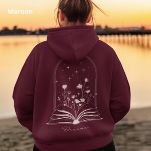 May include: A maroon hoodie featuring a white line art graphic on the back. The design depicts an open book with flowers growing out of the pages, enclosed within an arched frame with stars above. The word Dreamer is printed below the book.