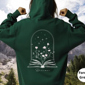May include: A forest green hooded sweatshirt featuring a white line art graphic on the back. The design depicts an open book with flowers growing from the pages inside an arched frame with stars. The word Dreamer is printed below the book.