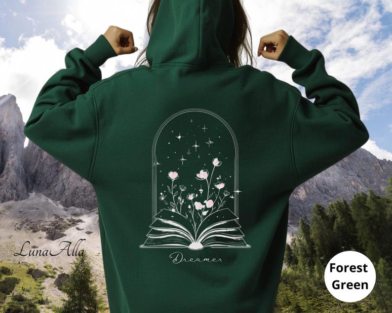 May include: A forest green hooded sweatshirt featuring a white line art graphic on the back. The design depicts an open book with flowers growing from the pages inside an arched frame with stars. The word Dreamer is printed below the book.