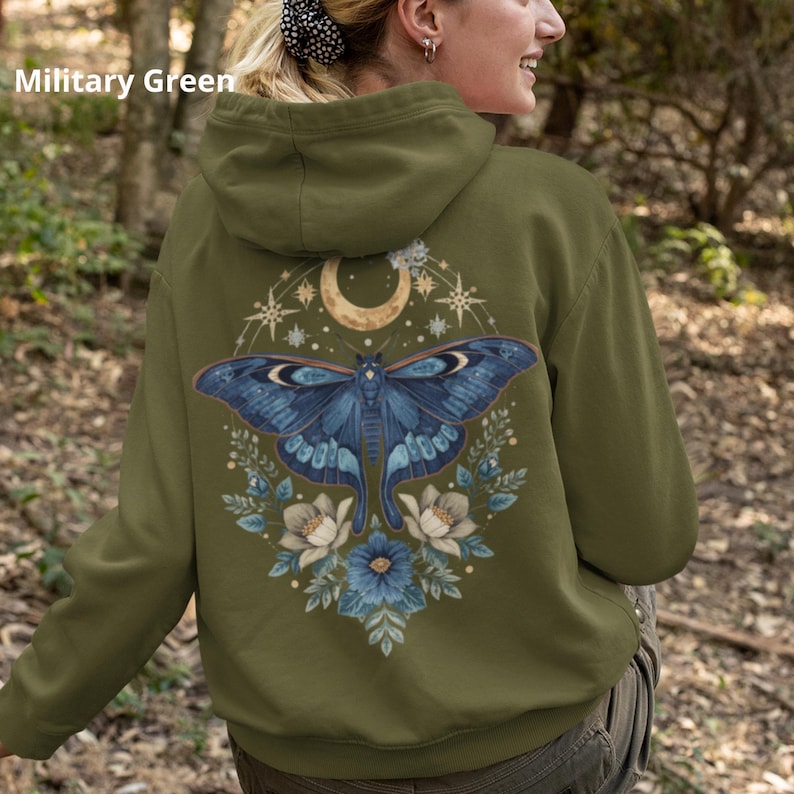 May include: A military green hooded sweatshirt featuring a large graphic on the back. The design displays a blue moth centered beneath a crescent moon and stars, surrounded by blue and white flowers and foliage. The text Military Green is visible.