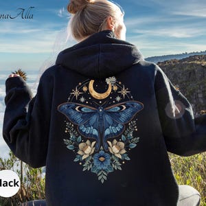 May include: A black hooded sweatshirt featuring a large graphic on the back. The design displays a blue moth centered beneath a golden crescent moon and stars, surrounded by blue and white floral elements. The text Luna Alla is visible.