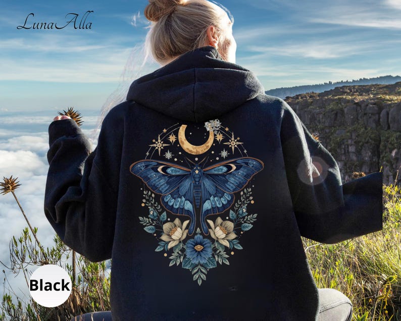 May include: A black hooded sweatshirt featuring a large graphic on the back. The design displays a blue moth centered beneath a golden crescent moon and stars, surrounded by blue and white floral elements. The text Luna Alla is visible.