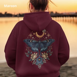 May include: A maroon hoodie featuring a back graphic of a large blue moth centered beneath a crescent moon, surrounded by starbursts and floral elements. The design includes intricate botanical details and celestial motifs in muted tones.