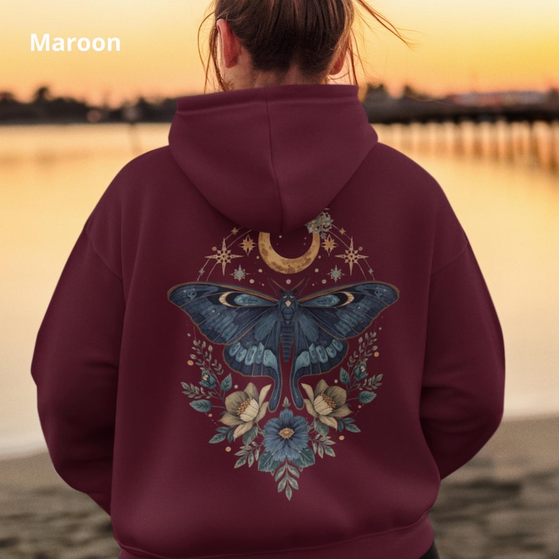 May include: A maroon hoodie featuring a back graphic of a large blue moth centered beneath a crescent moon, surrounded by starbursts and floral elements. The design includes intricate botanical details and celestial motifs in muted tones.