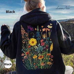 May include: A black hooded sweatshirt featuring a large, colorful graphic on the back. The print displays a variety of wildflowers in shades of yellow, orange, blue, and purple. The text "Luna Alla" is printed in the upper right corner of the image.