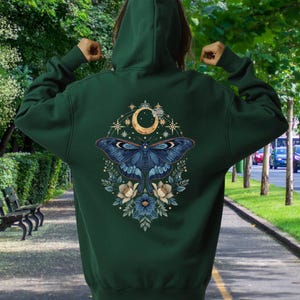 May include: A dark forest green hooded sweatshirt featuring a large back graphic of a blue moth centered beneath a golden crescent moon, surrounded by intricate star patterns and floral elements in shades of blue, cream, and gold.