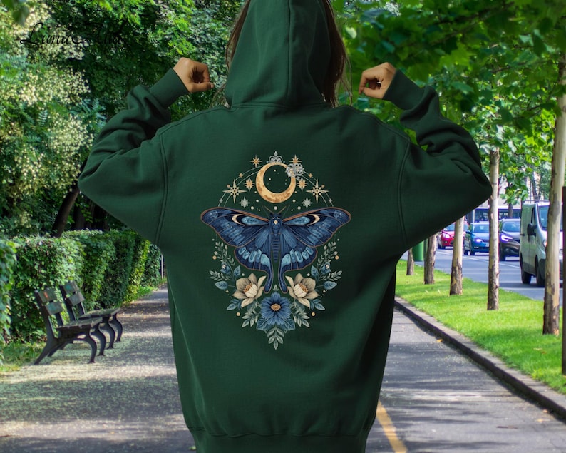 May include: A dark forest green hooded sweatshirt featuring a large back graphic of a blue moth centered beneath a golden crescent moon, surrounded by intricate star patterns and floral elements in shades of blue, cream, and gold.